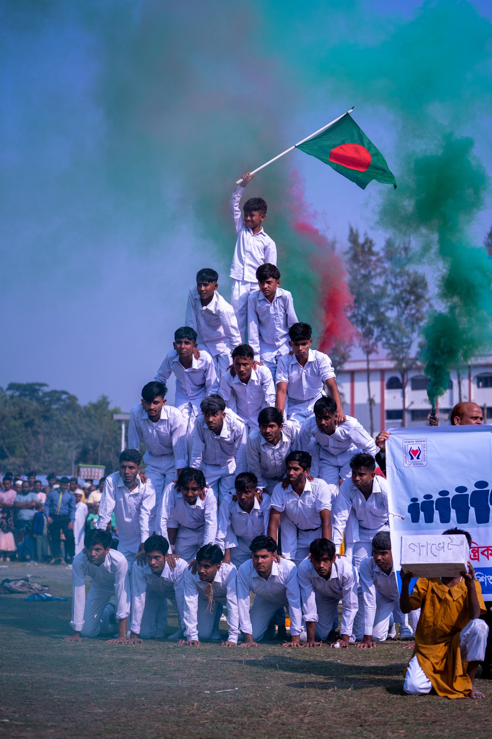 Boys in white forming a human pyramid with one atop waving the Bangladesh flag amid red and green smoke