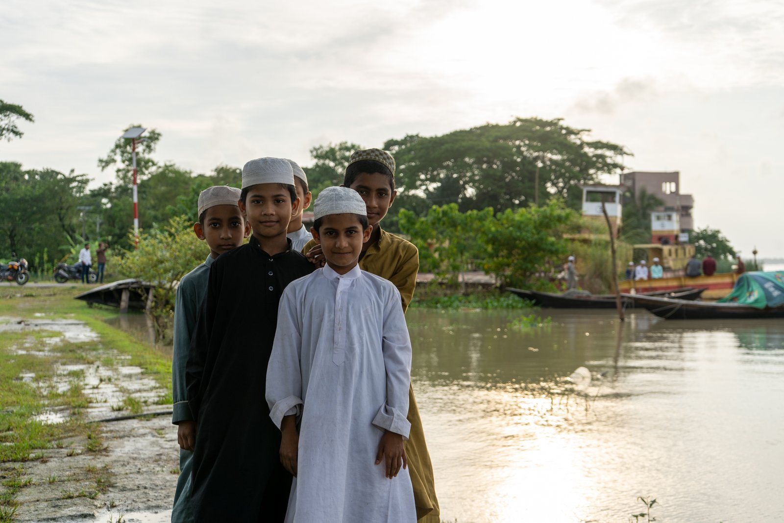 Four boys in traditional dress standing together by a river at sunset near a wooden dock
