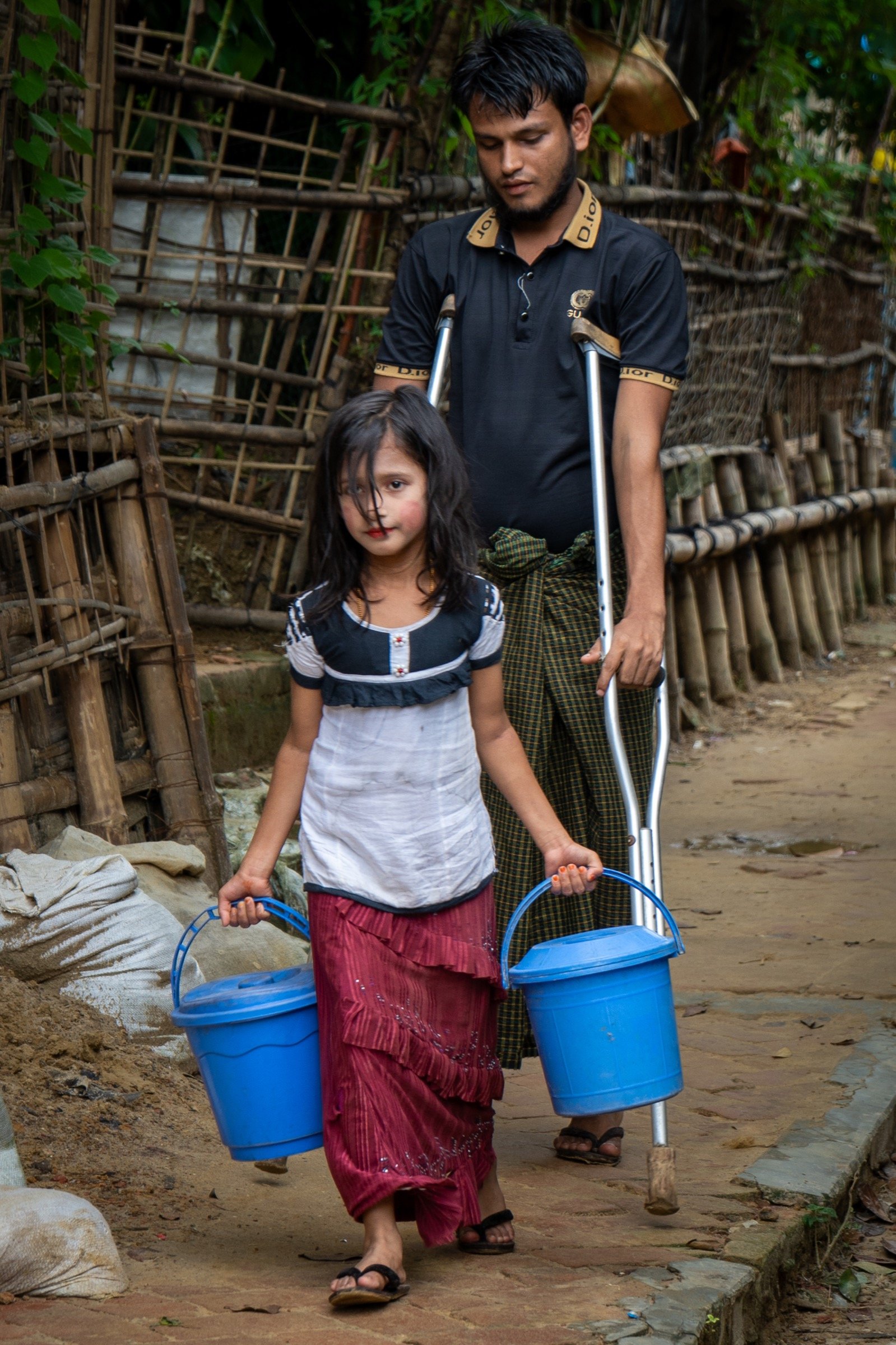 A man on crutches and a child with blue buckets near a camp water point