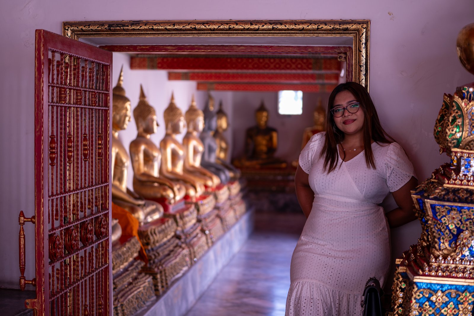 Woman in a white lace dress standing at the entrance of a gilded Buddhist statue corridor, framed by an ornate doorway