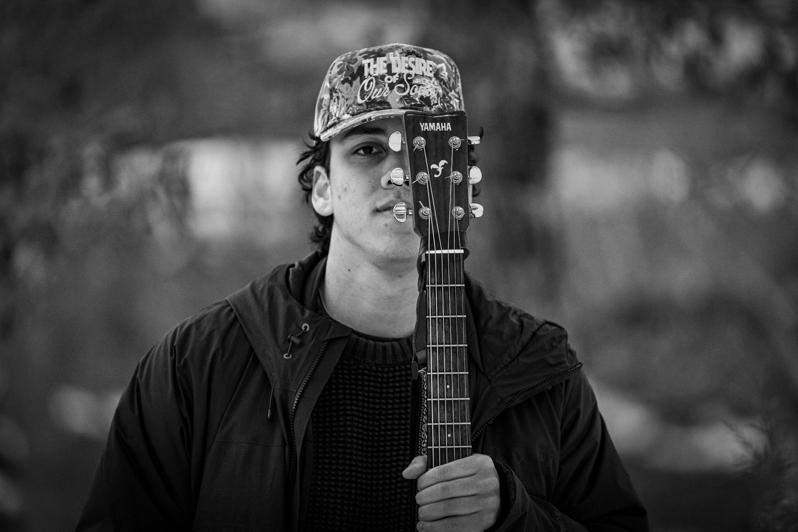 Young man in a patterned cap holding a Yamaha guitar neck across his face in a black-and-white close portrait