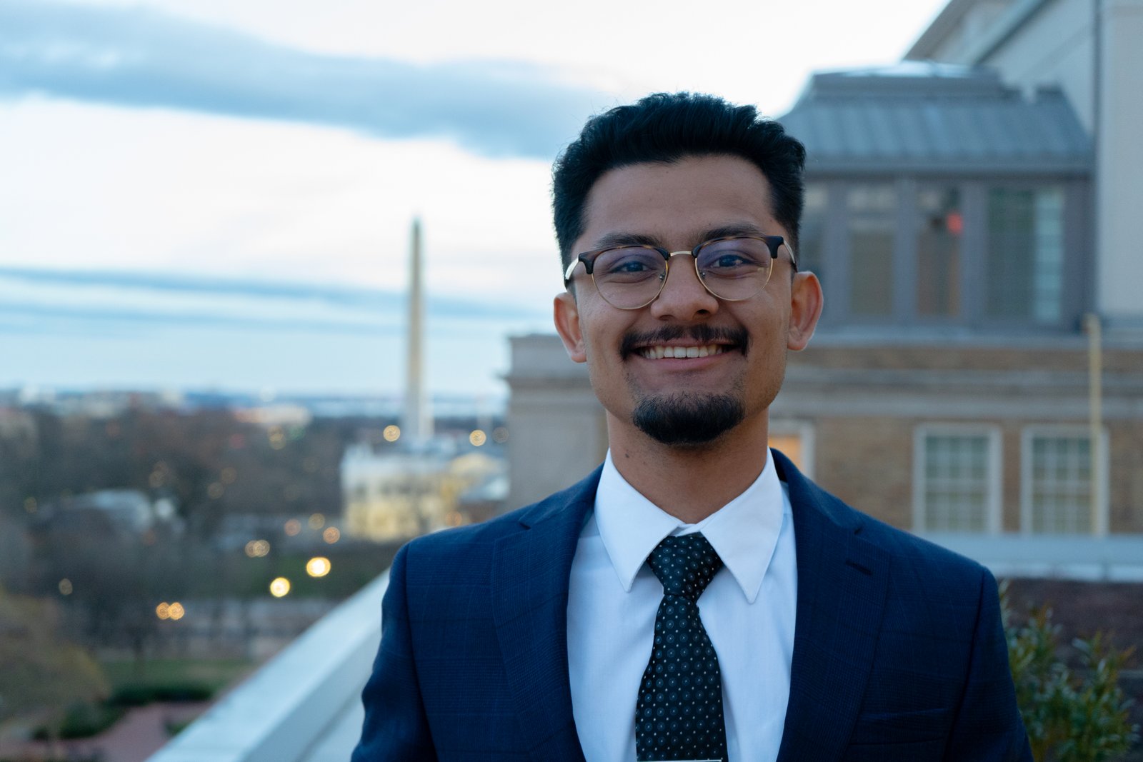 Young man in a navy suit and tie smiling on a rooftop, the Washington Monument visible in the soft-focus background