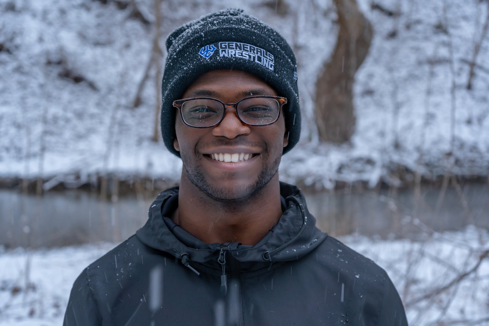 Young man smiling broadly while wearing a wrestling team beanie with snow falling around him