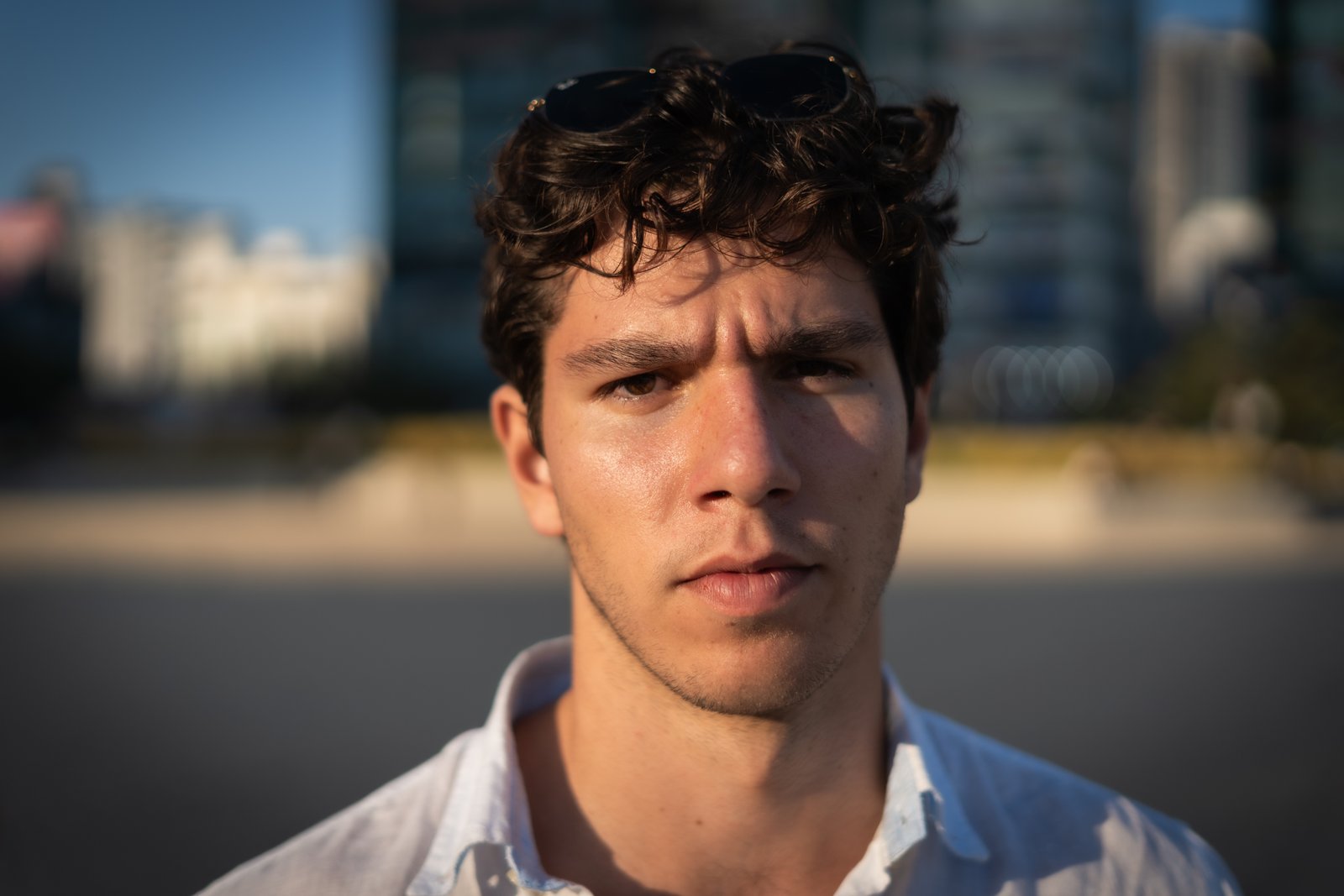 Young man with curly hair and sunglasses pushed up on his head, direct gaze, city buildings softly out of focus behind him