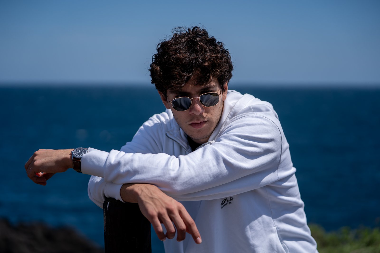 Young man in a white hoodie and aviator sunglasses leaning on a railing with a deep blue ocean horizon behind him